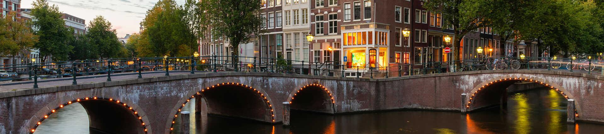 Amsterdamer Kanal bei Dämmerung, mit spiegelnden Lichtern im Wasser und traditionellen niederländischen Häusern mit Giebeln am Ufer.