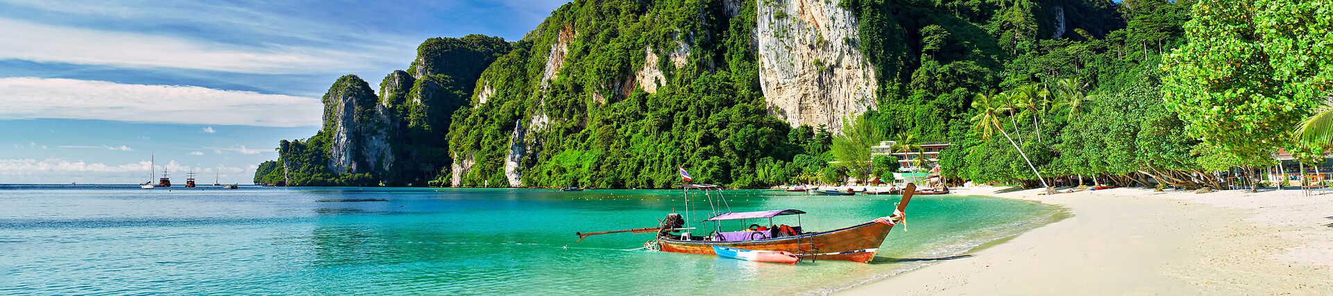Weißer Sandstrand auf den Phi Phi Inseln mit einem traditionellen Holz-Langheckboot im klaren türkisfarbenen Meer und üppig bewachsenen Kalksteinfelsen im Hintergrund.