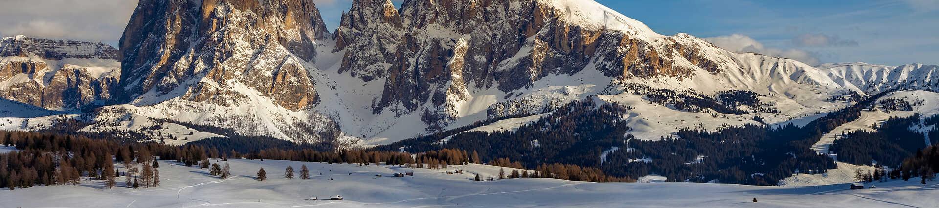 Weite, schneebedeckte Landschaft der Seiser Alm in Südtirol, mit Blick auf die markanten, zerklüfteten Felsgipfel der Langkofelgruppe (Langkofel und Plattkofel) im Winter. Teil des UNESCO-Welterbes.