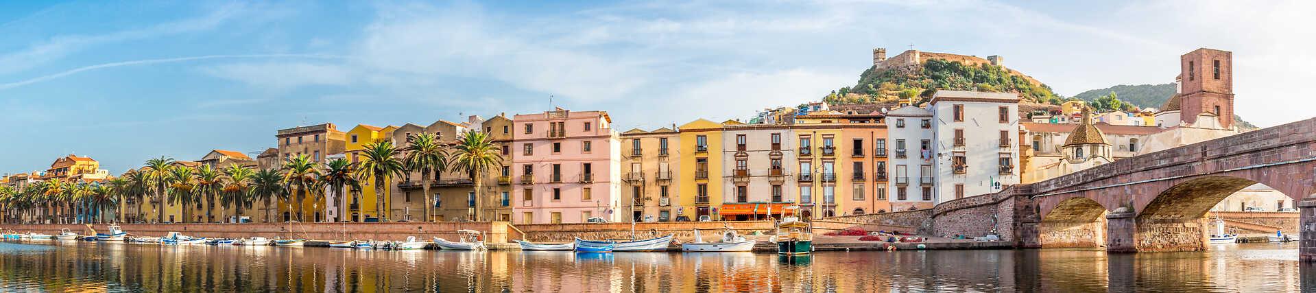 Panoramabild der charmanten Stadt Bosa in Italien, mit pastellfarbenen Häusern, die sich am Ufer des einzigen schiffbaren Flusses Sardiniens, dem Temo, spiegeln, sowie der alten Ponte Vecchio Brücke und dem mittelalterlichen Schloss auf dem Hügel im Hintergrund.