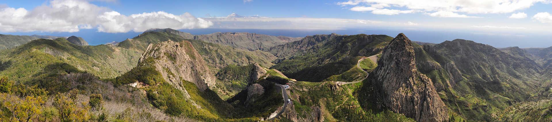 Panoramablick über eine grüne, zerklüftete Berglandschaft mit tiefen Schluchten; eine kurvenreiche Straße schlängelt sich über einen schmalen Bergrücken. Im Hintergrund erstrecken sich das blaue Meer und ein wolkiger Himmel über dem Horizont.