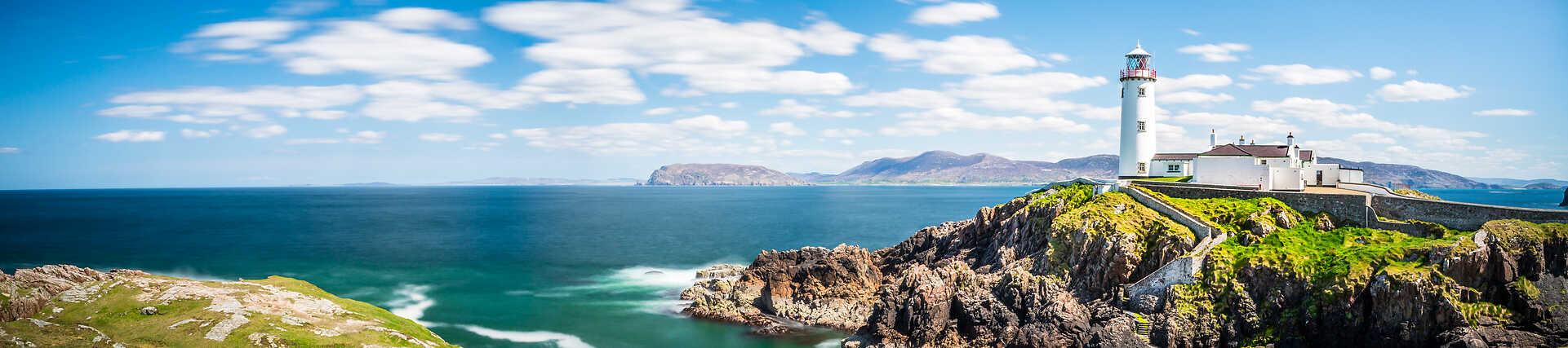 Panorama-Luftaufnahme des weißen Fanad Head Lighthouse mit roter Kappe auf einer felsigen Klippe, umgeben von der wilden Küste Irlands und dem blauen Atlantik. Teil des Wild Atlantic Way.