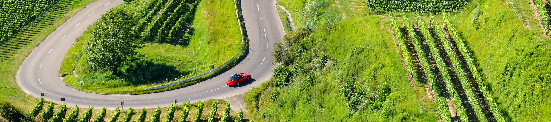 Rotes Cabrio auf kurviger Straße im Kaiserstuhl zwischen grünen Weinbergen.
