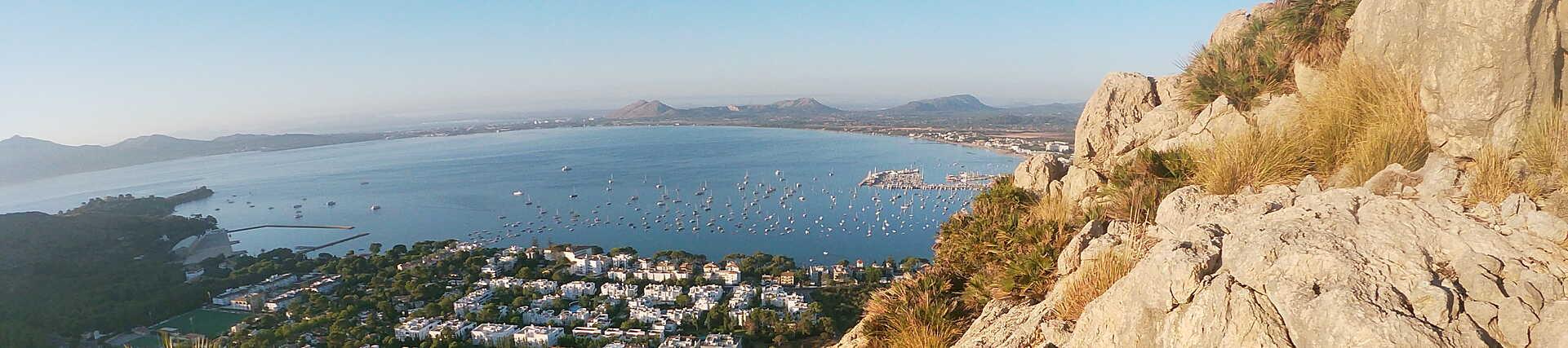 Panorama-Aufnahme aus der Vogelperspektive, aufgenommen von den umliegenden Klippen oder Bergen, die die gesamte Bucht von Pollença auf Mallorca in Spanien zeigt.