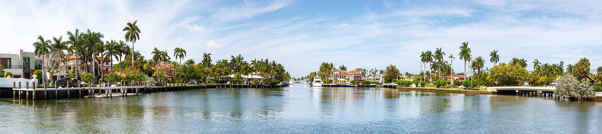 Panorama eines breiten Kanals in Fort Lauderdale, gesäumt von luxuriösen, mediterran inspirierten Villen mit Gärten, Palmen und privaten Bootsanlegestellen an einem sonnigen Tag.