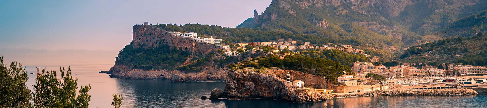Panorama-Luftaufnahme der geschützten Bucht von Port de Sóller auf Mallorca, mit dem Far de sa Creu Leuchtturm auf der Klippe, dem Hafen und der Stadt, umgeben von grünen Bergen.