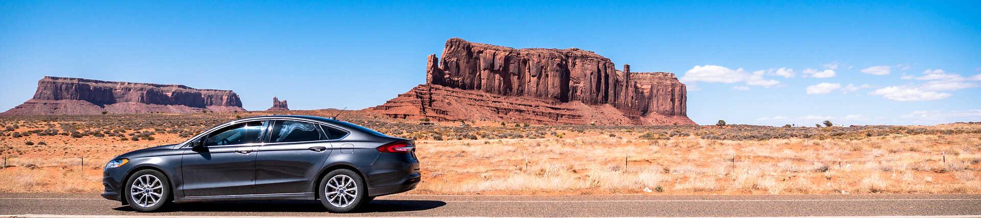 Mietwagen fährt auf der US-Highway 163 durch die ikonische Monument Valley Wüstenlandschaft mit großen roten Sandstein-Felsformationen in der Ferne.