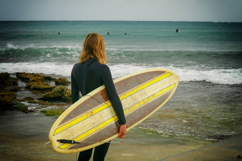 Frau mit Surfbrett steht an einem Sandstrand und schaut auf das Wasser